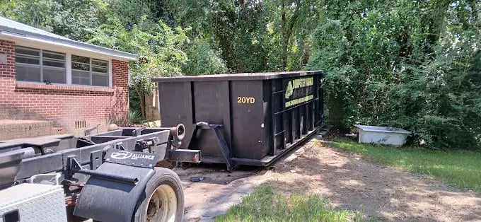 Large dumpster and trailer parked beside brick building with trees
