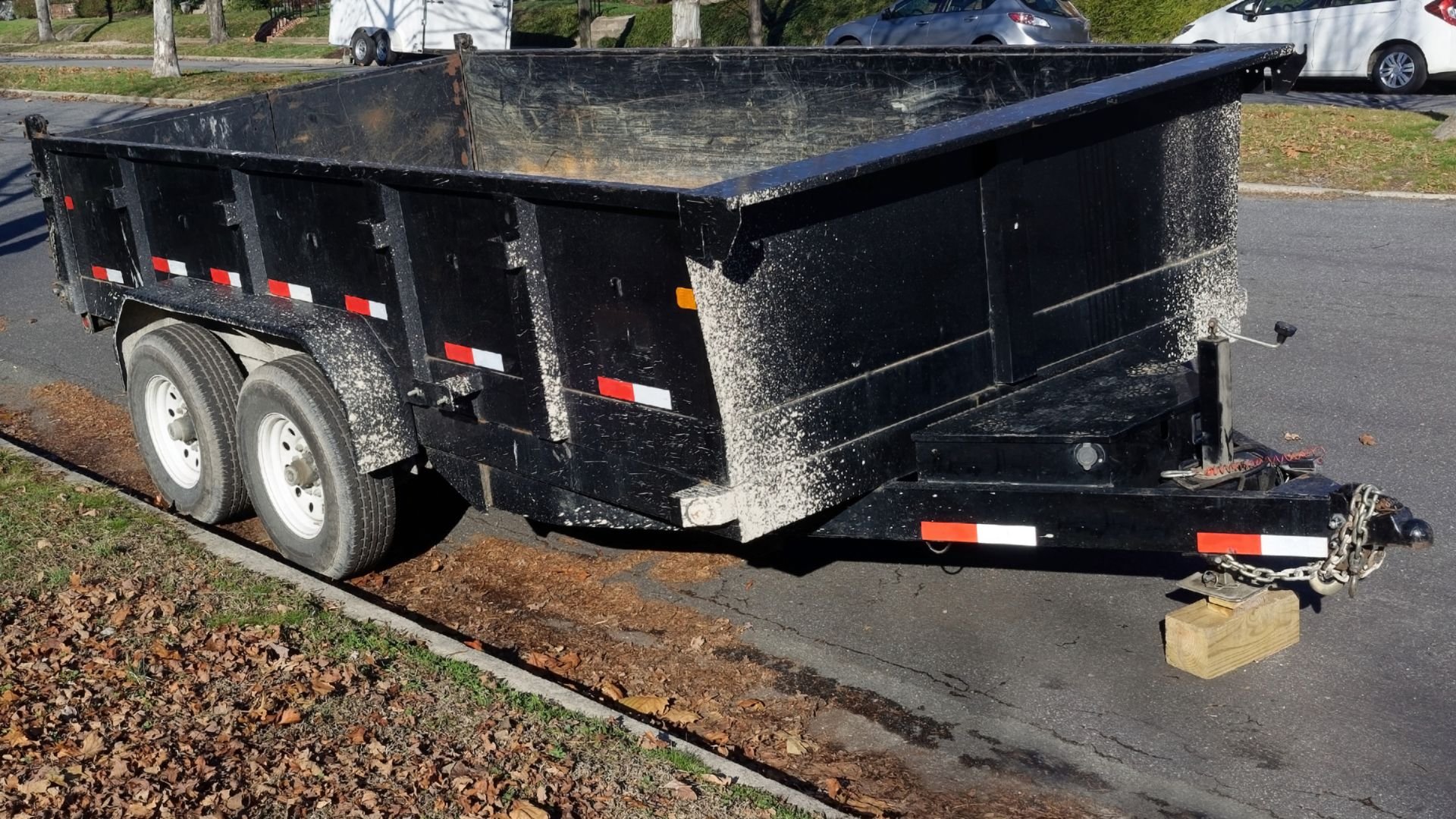 Black two-axle dump trailer parked on street with fallen leaves