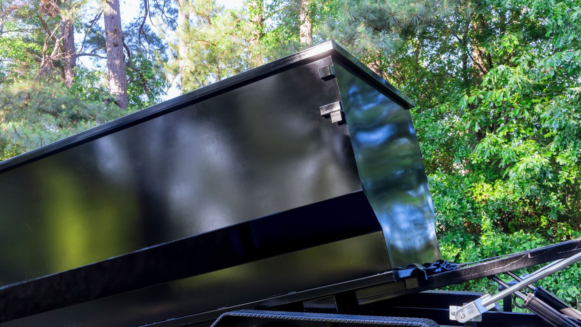 Black dumpster tilted open in a forested area with green trees