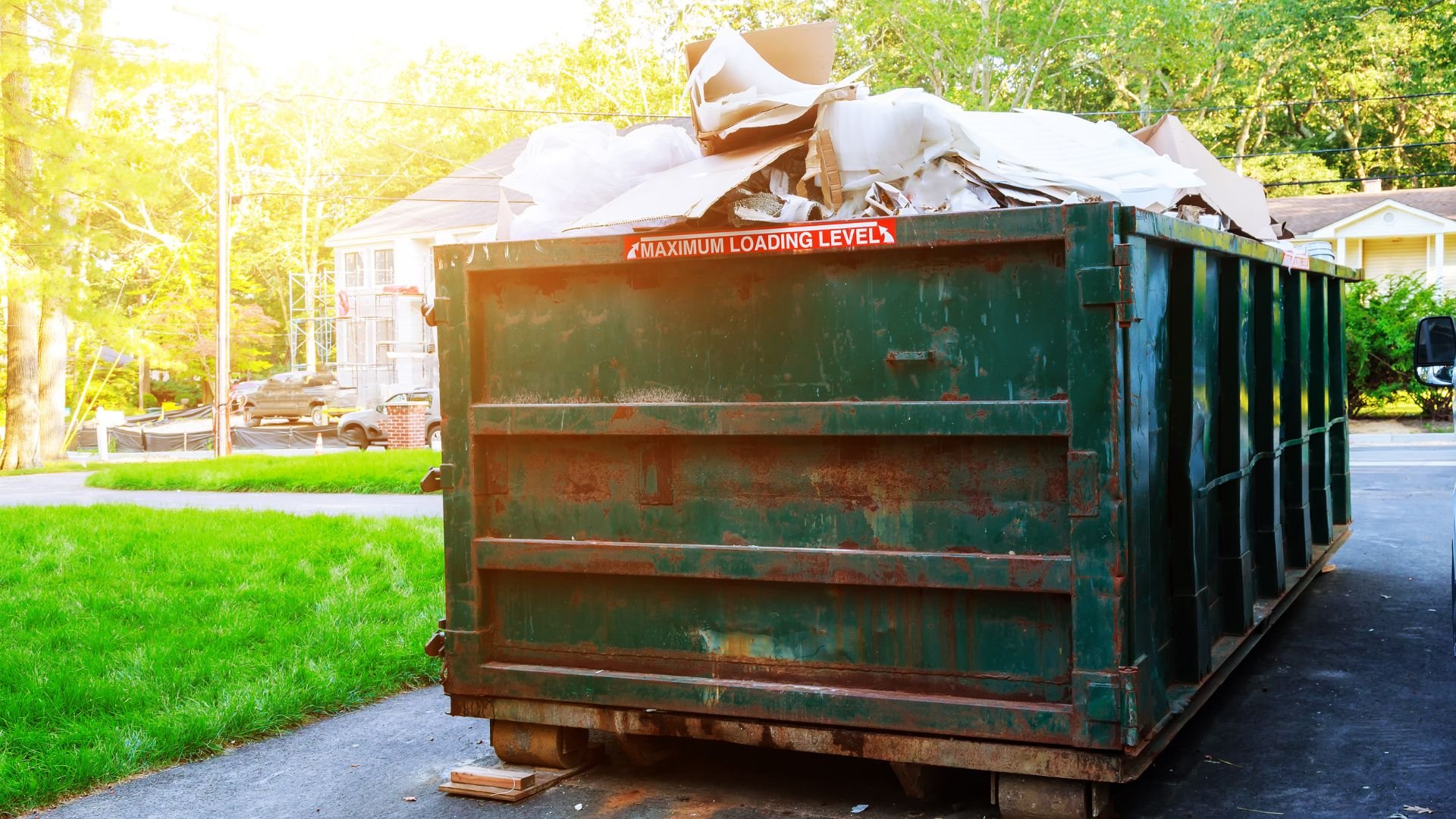 Overflowing green dumpster with debris on residential street in sunlight